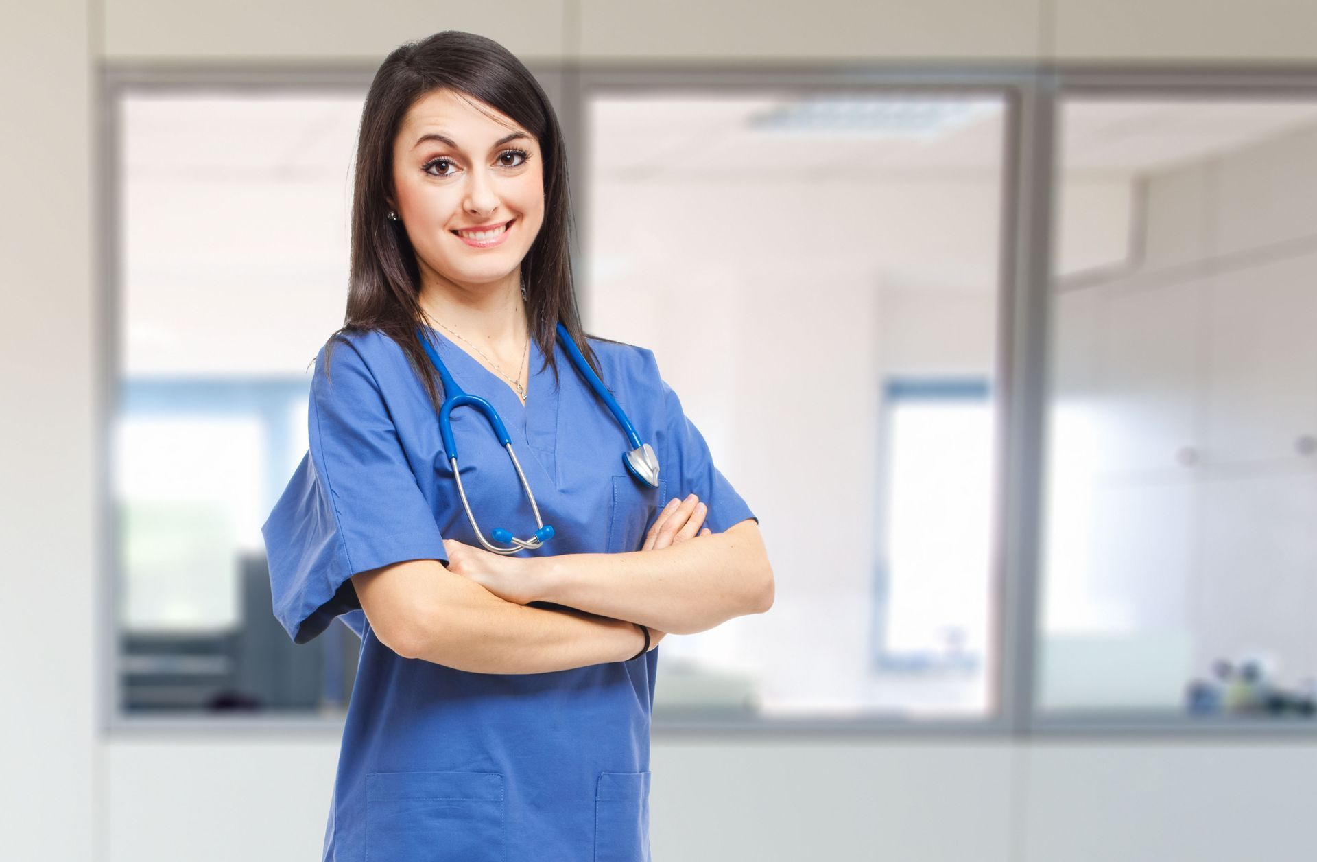 A healthcare professional in blue scrubs with a stethoscope around their neck, smiling with arms crossed in an office.