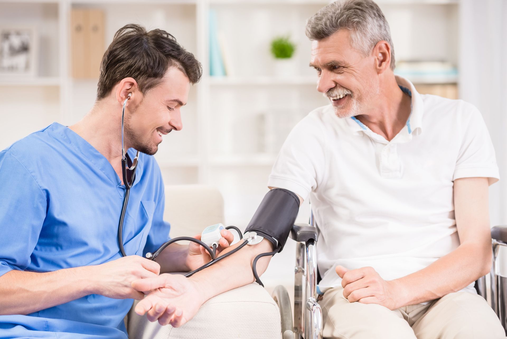 A healthcare worker in blue scrubs checks a patient's blood pressure with a cuff and stethoscope in a clinical setting.