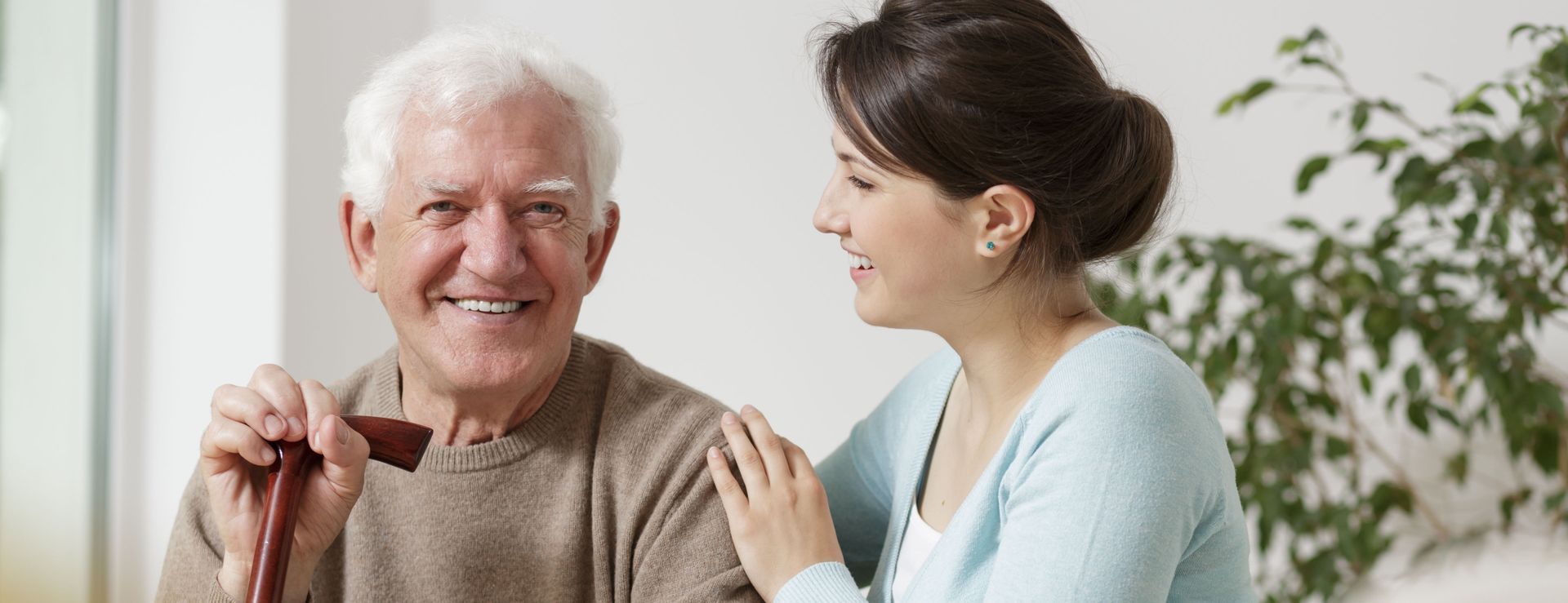 A person holding a cane smiles at a caregiver who rests a hand on their shoulder in a bright, indoor setting.
