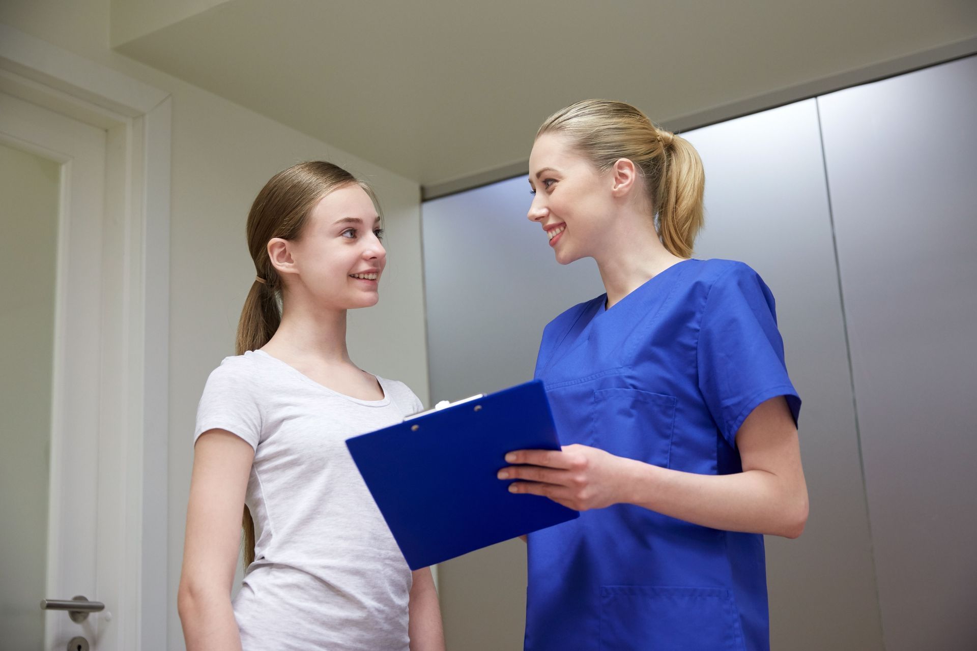 A healthcare professional in a blue scrub top talks to a young person while holding a blue clipboard.