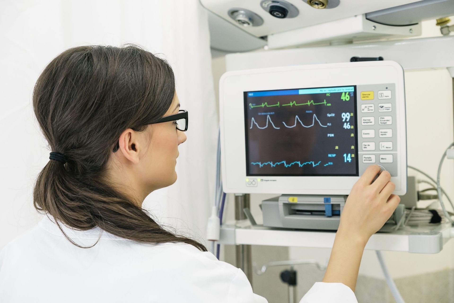A medical professional in a white coat adjusts settings on a patient bedside monitor in a clinical setting.