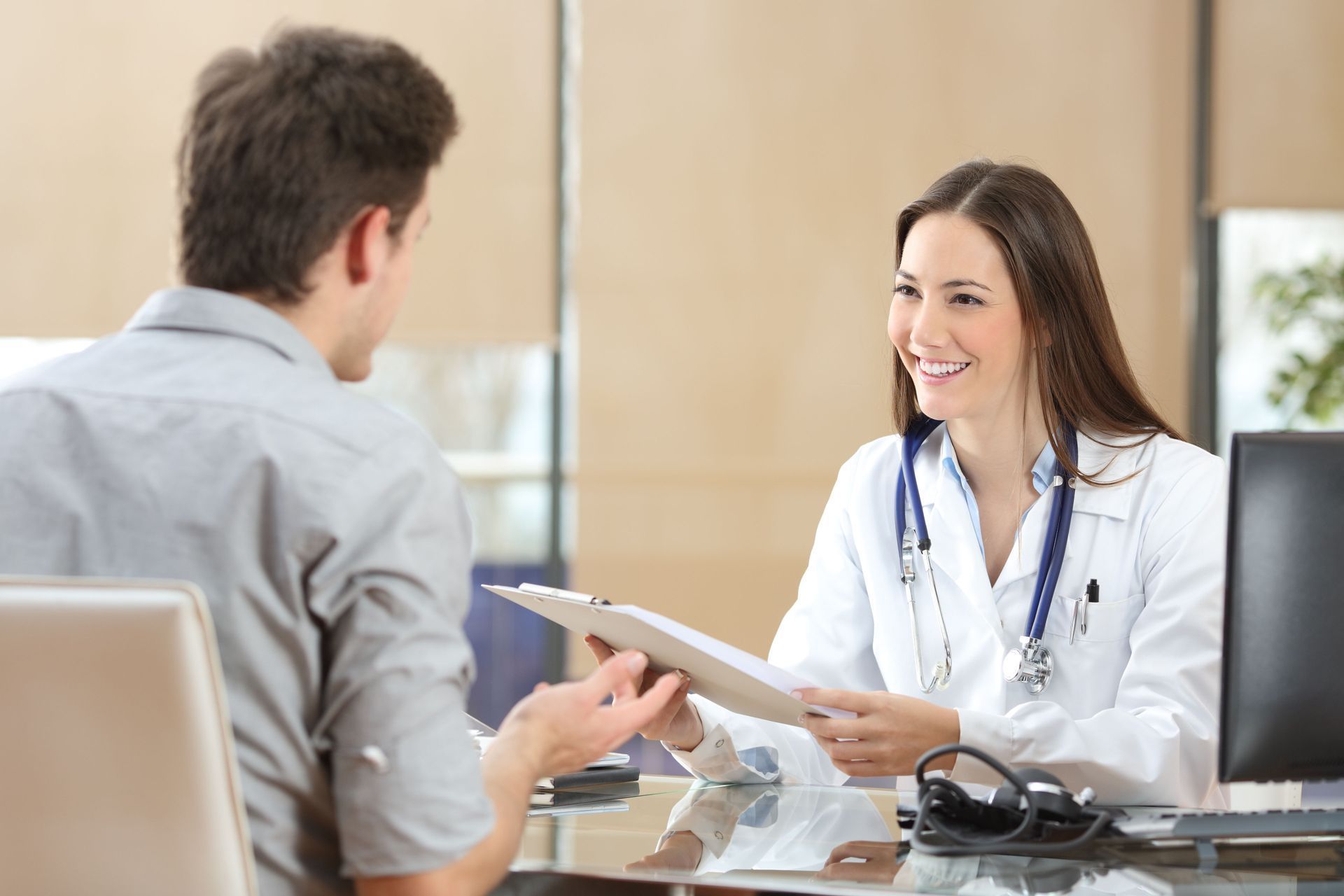 A smiling doctor in a white coat with a stethoscope holds a clipboard, talking to a patient in an office setting.