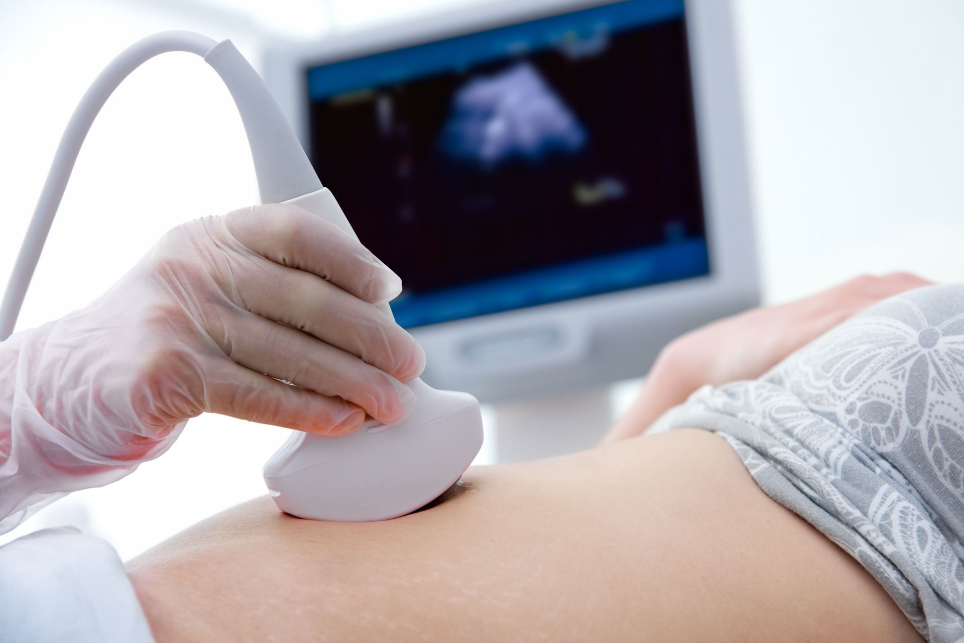 A person in a medical glove uses an ultrasound transducer on a patient's abdomen, with a monitor displaying a scan.