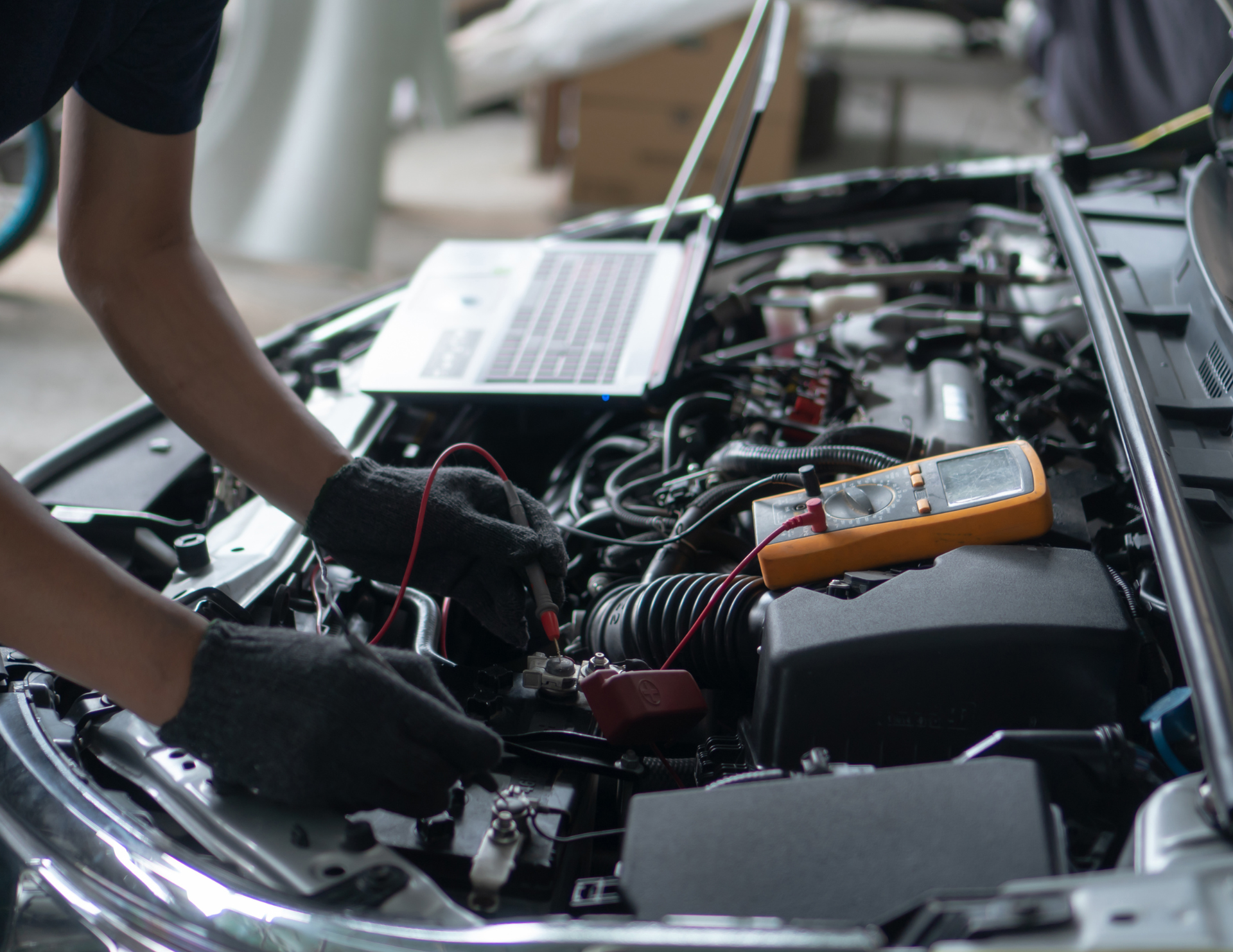 Mechanic working on car engine, using laptop and multimeter.
