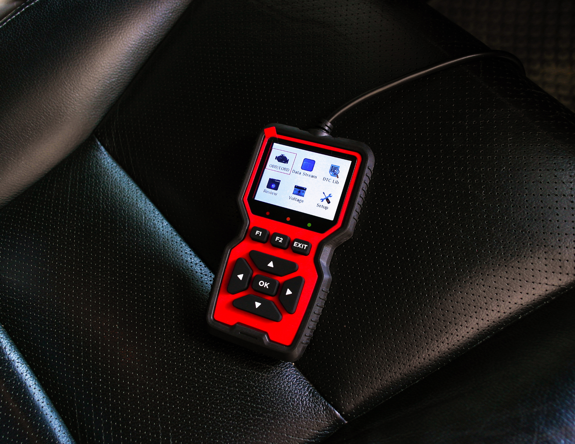 Red and black OBD2 scanner on black leather car seat, display showing icons, attached cord.