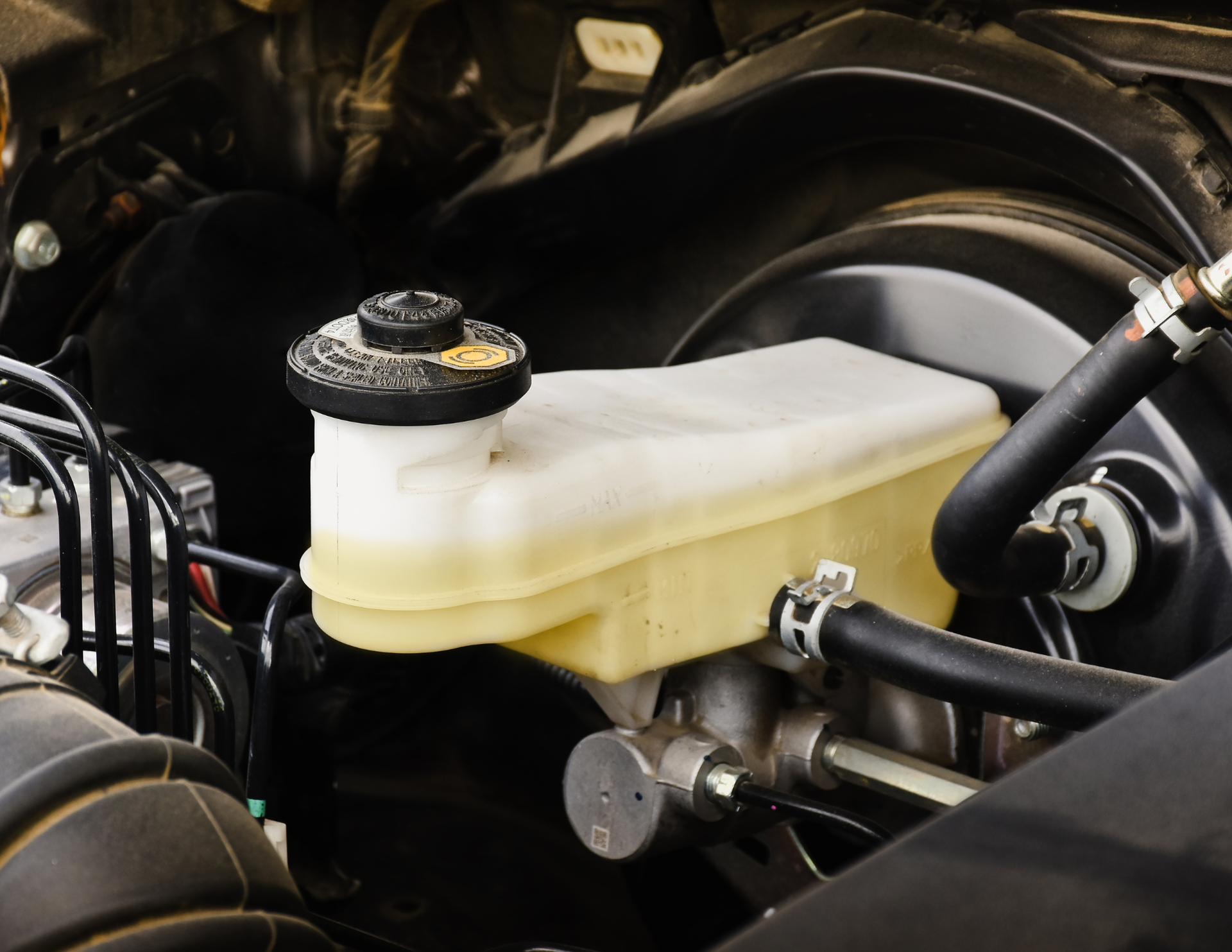 Brake fluid reservoir in a car engine bay, yellow fluid visible, black cap and hoses.