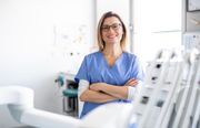 A female dentist is standing in a dental office with her arms crossed.