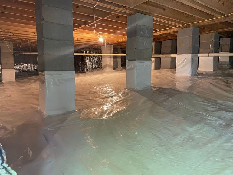 Crawlspace with concrete block supports wrapped in white, covered by plastic sheeting. Wooden beams above.