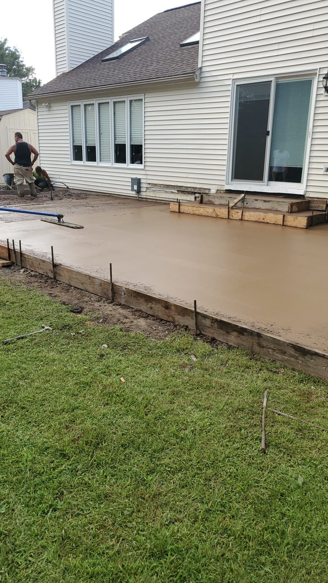 Concrete patio being poured next to a house with a person standing nearby. Green grass in foreground.