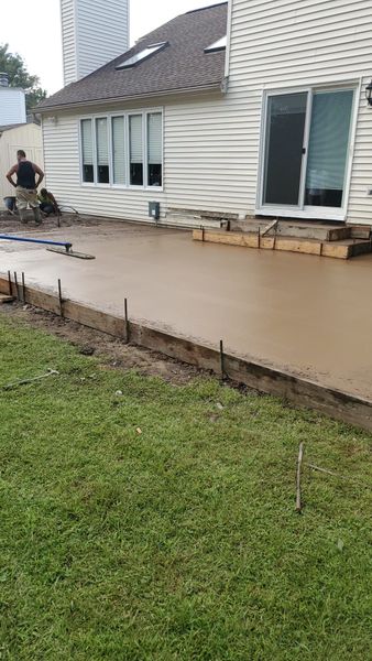 Concrete patio being poured next to a house with a person standing nearby. Green grass in foreground.