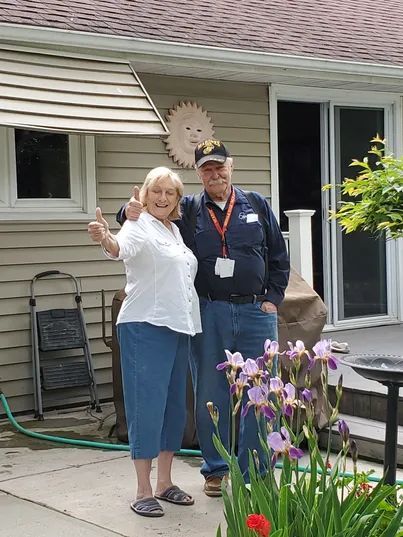 Woman giving thumbs up, smiling, with man in front of a house, surrounded by plants and outdoor decor.