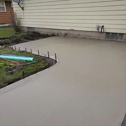 Freshly poured concrete patio next to a beige-sided house. Green grass and construction materials are visible.