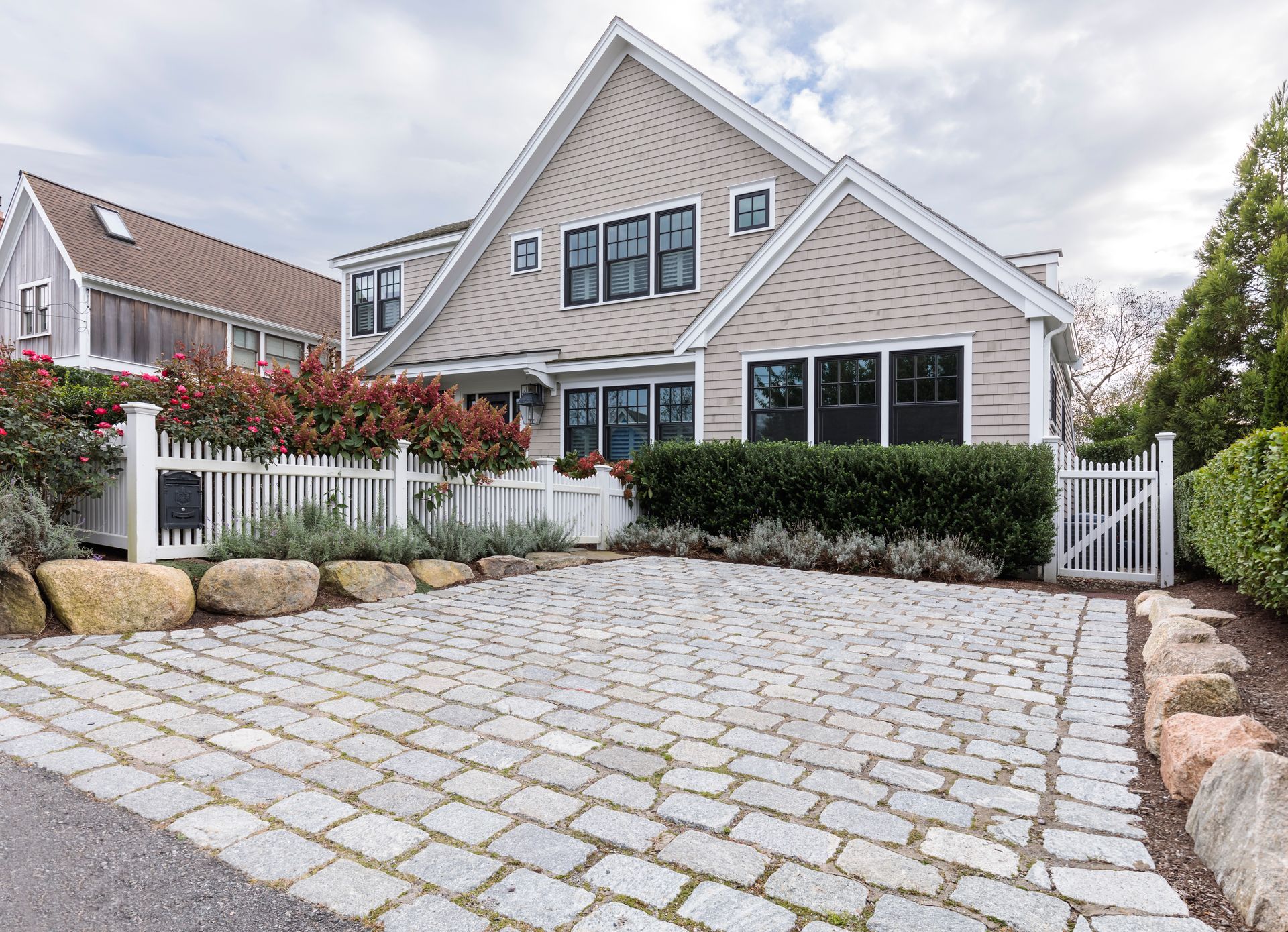 Beige and gray house with cobblestone driveway, white picket fence, and green hedges.