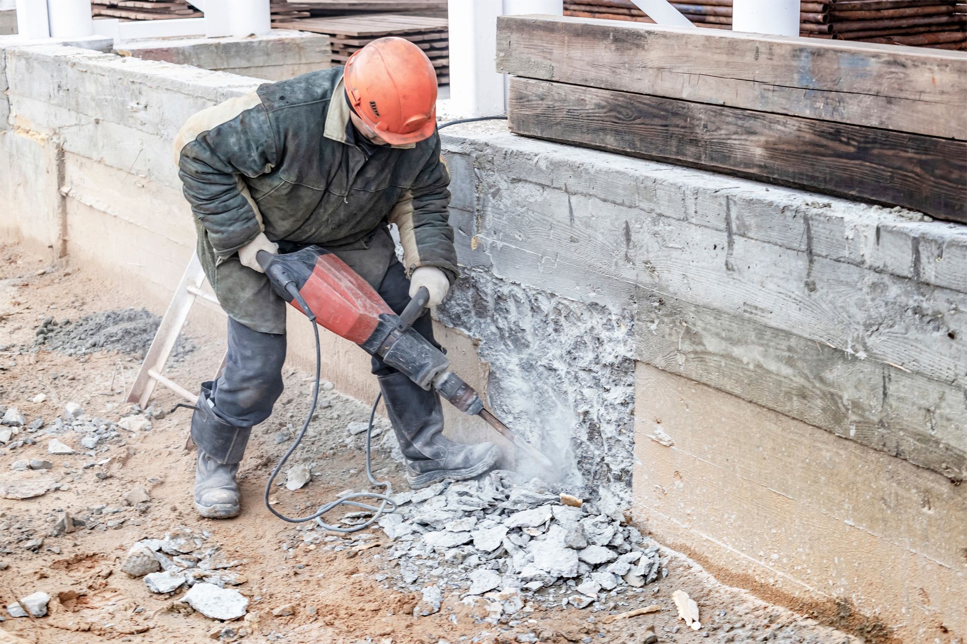Construction worker using a jackhammer to break concrete outdoors. He wears a helmet and work clothes.
