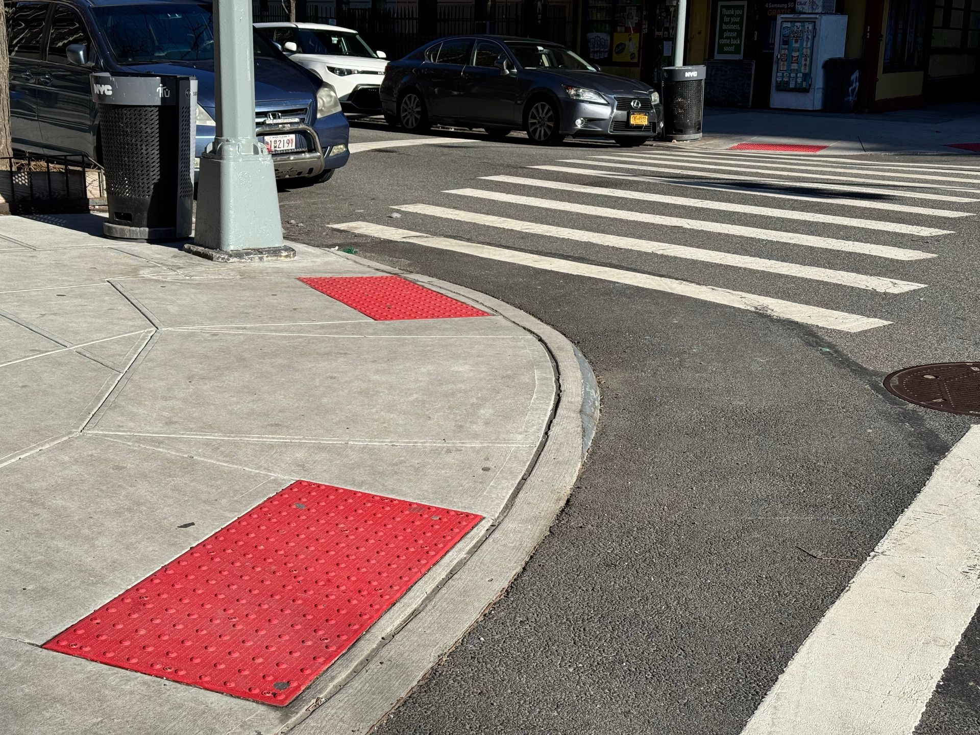 Sidewalk corner with red tactile paving, crosswalk, parked cars, and street in daylight.