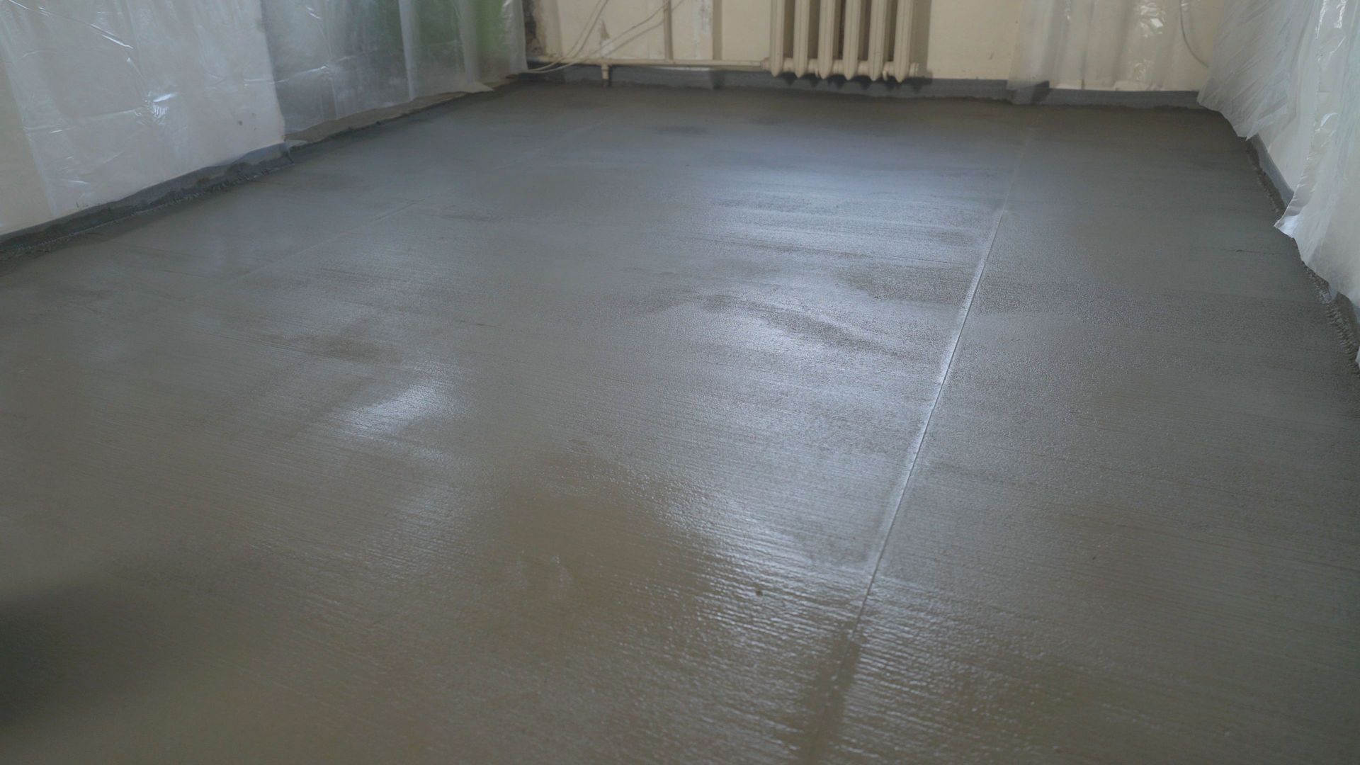 Newly poured, smooth gray concrete floor in a room, with plastic sheeting on walls.