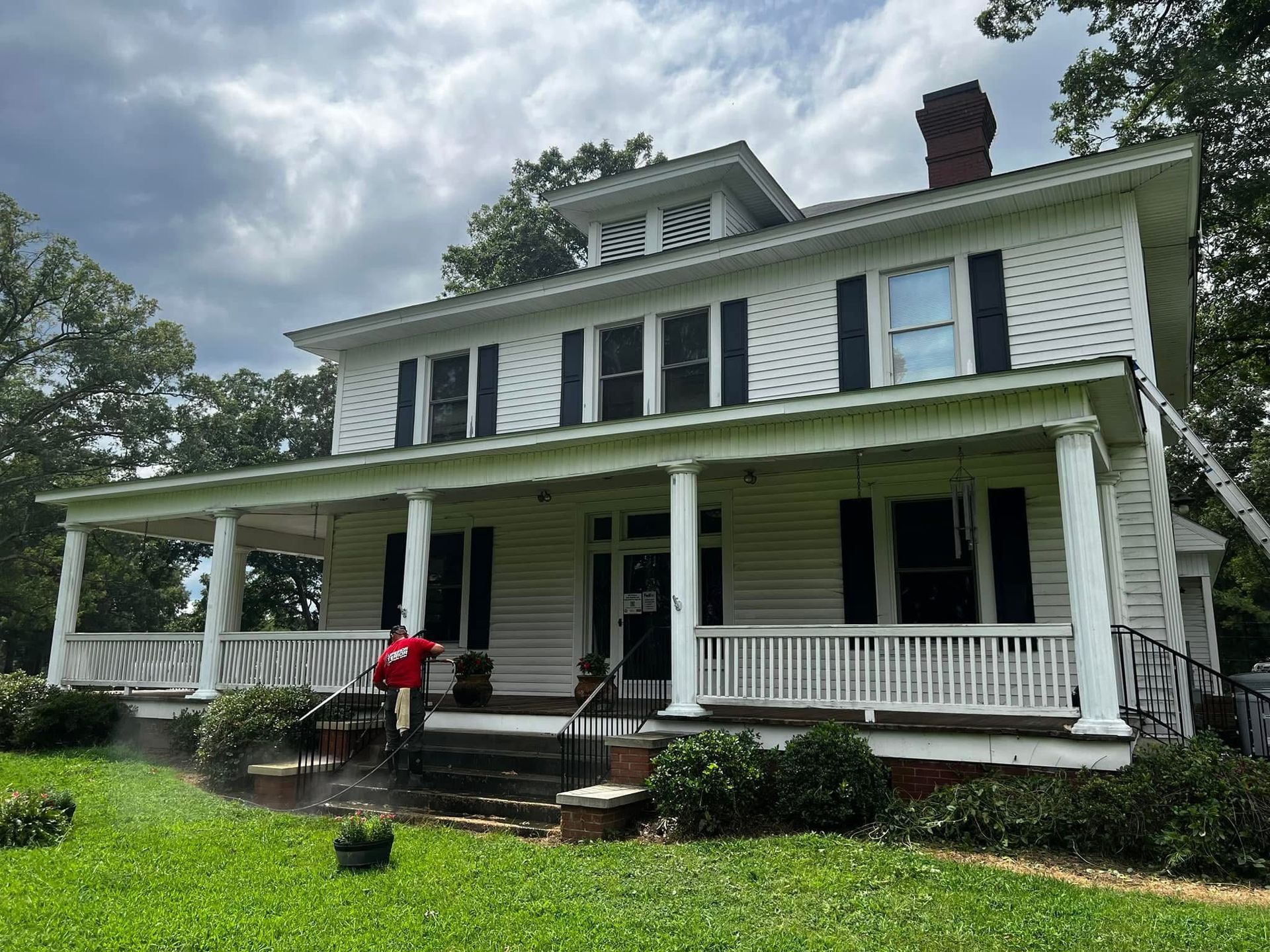A man in a red shirt is standing on the porch of a large white house.