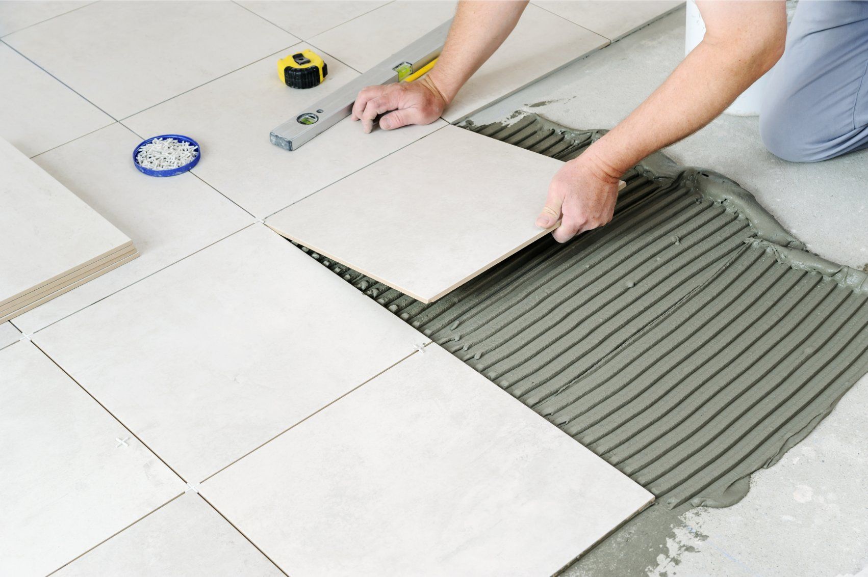 Man laying light-colored tile on a floor, using a level and a trowel to apply mortar.