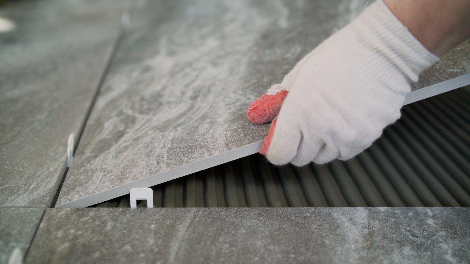 Gloved hand placing a gray tile next to other tiles on a surface with mortar and spacers.