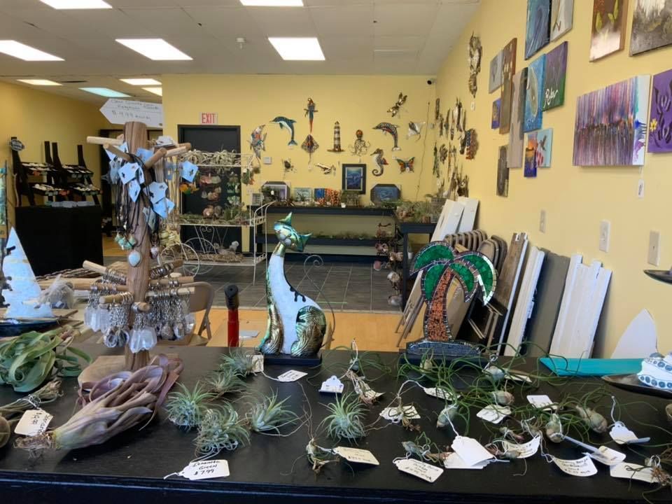 A retail shop with artistic displays. Features a table with plants, jewelry, and wall art against a yellow wall.