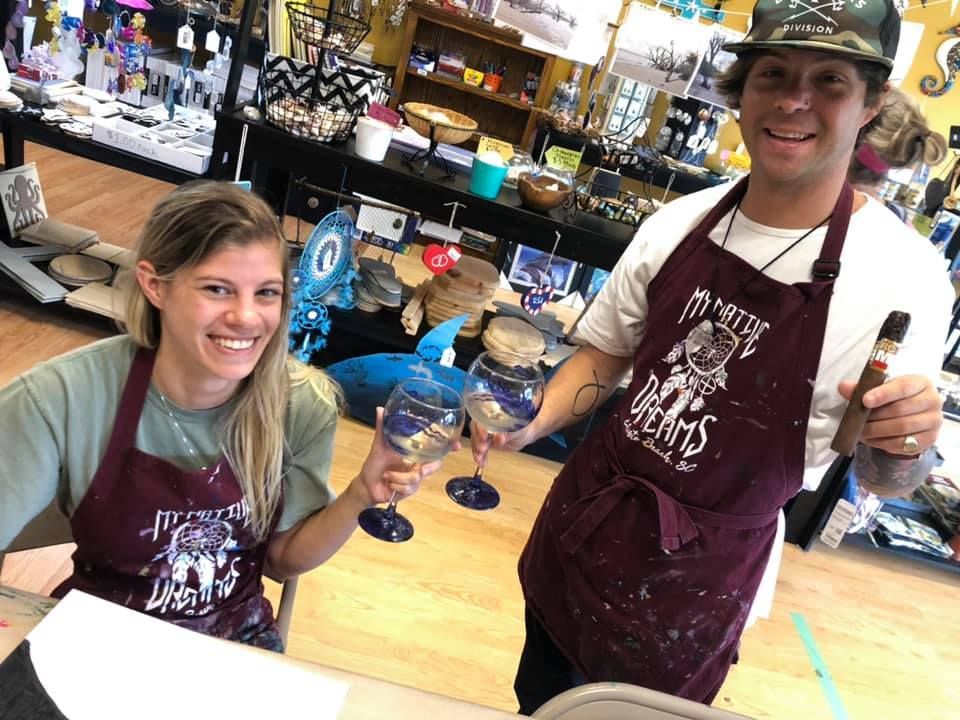 Smiling couple in aprons with painted wine glasses, man holding cigar in shop in a USA shop.