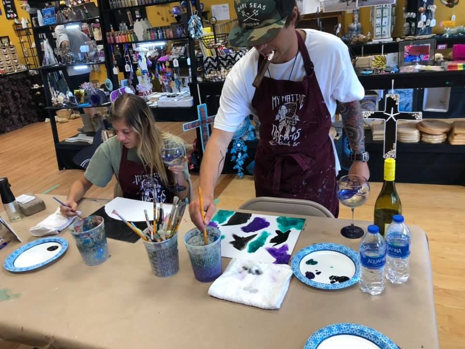 Two people painting at a table, using paint and brushes. Wine and water bottles  in a USA shop.are present.