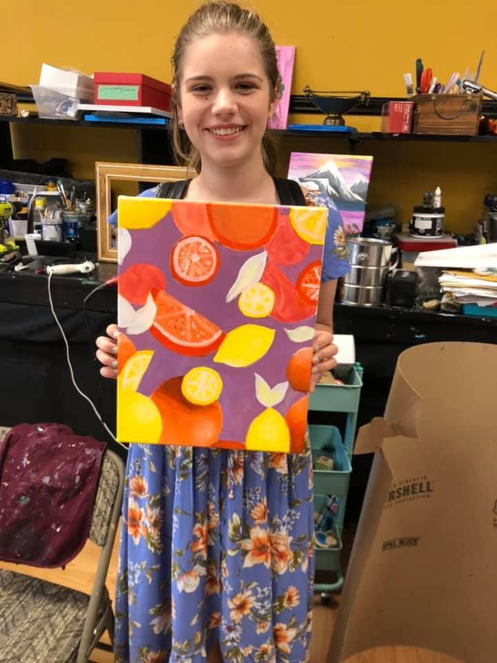 Girl holding a citrus fruit painting, smiling. Purple background, oranges, lemons, grapefruit in colorful shapes in a USA shop.