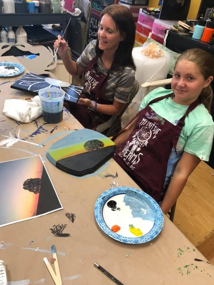 Two women painting together at a table, smiling. One holds a brush, the other has a palette in a USA shop.