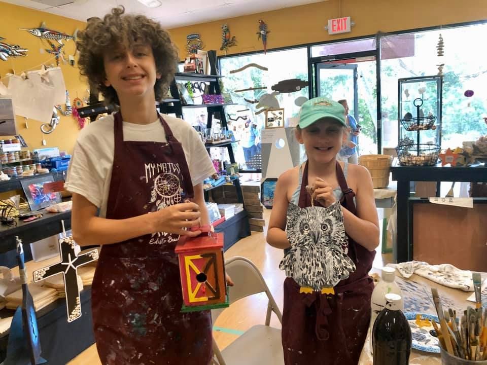 Two kids in aprons smile, holding painted birdhouses in an art studio in a USA shop.