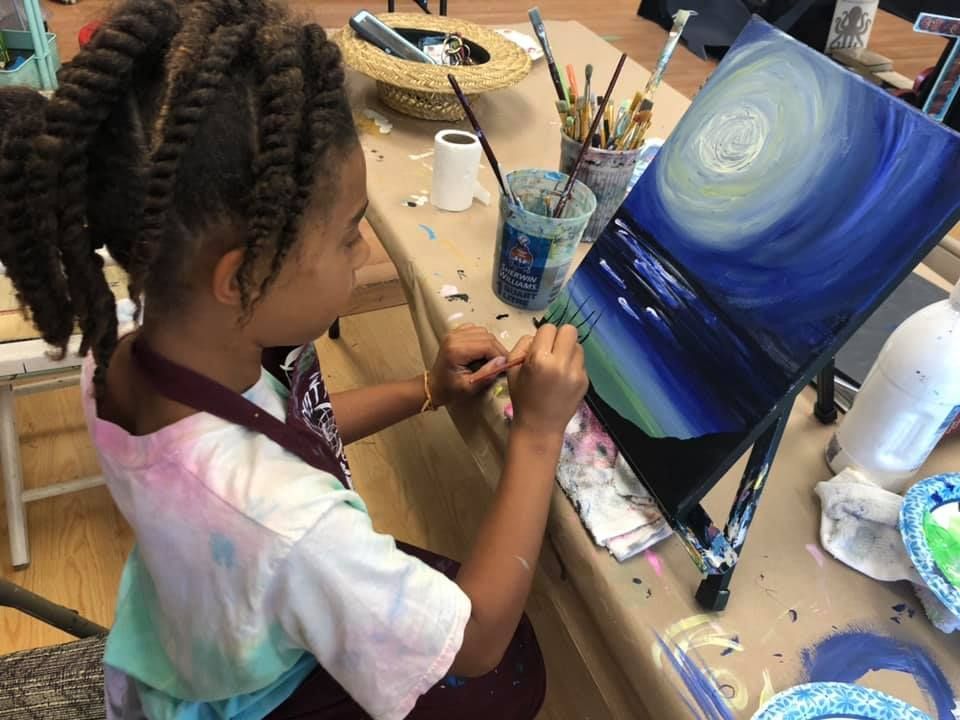 Girl with braided hair painting a blue and white landscape on a canvas at a table in a studio in a USA shop.