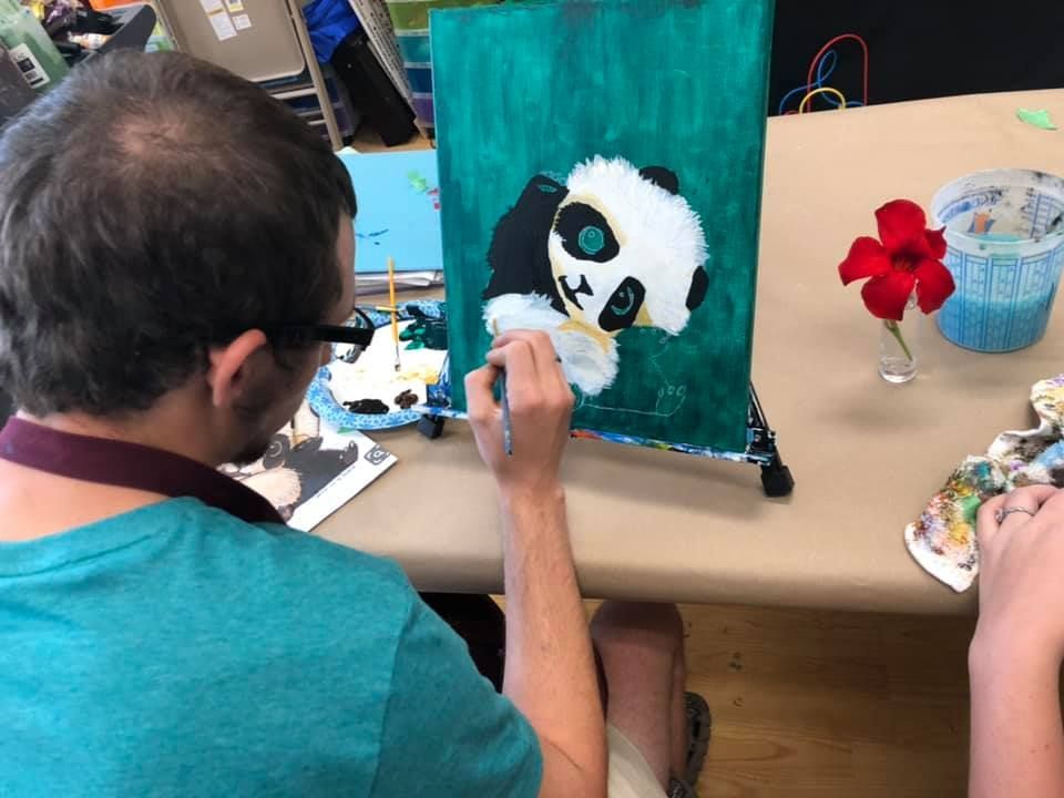 Person painting a panda on a canvas; green background; red flower in a vase in a USA shop.