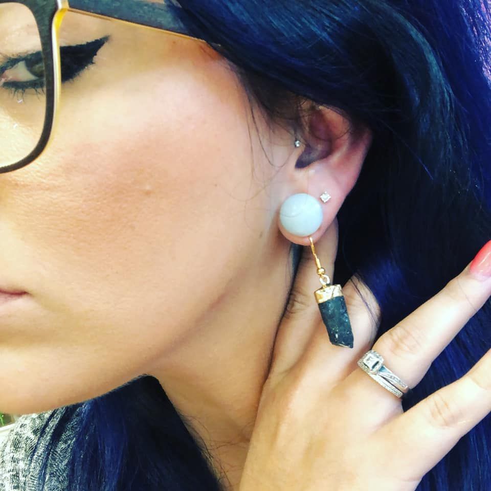 Woman with dark blue hair and glasses, wearing a large white earring and a stone earring.  She's touching her jaw, with a ring in a USA shop.