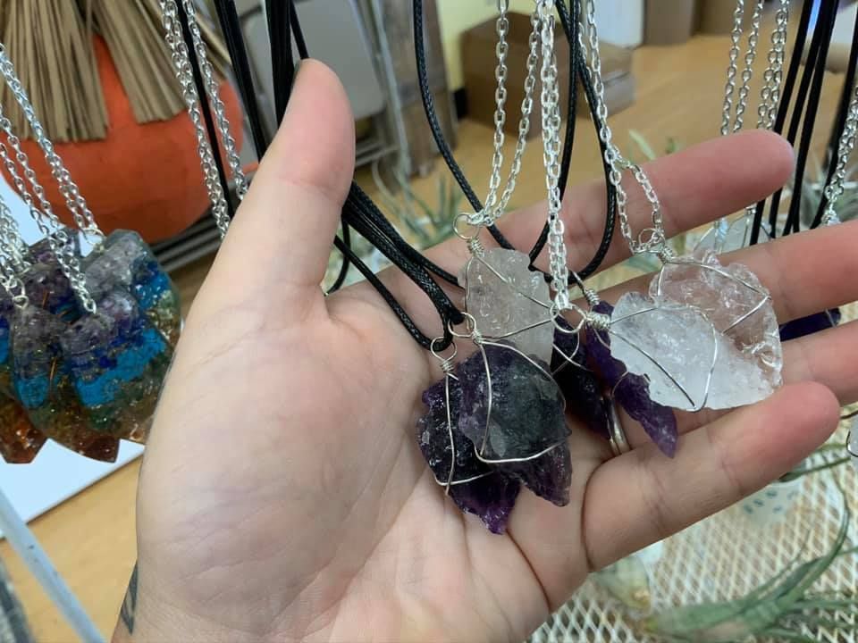 Hand holding several crystal pendant necklaces, featuring various colors and shapes, some wired in silver in a USA shop.