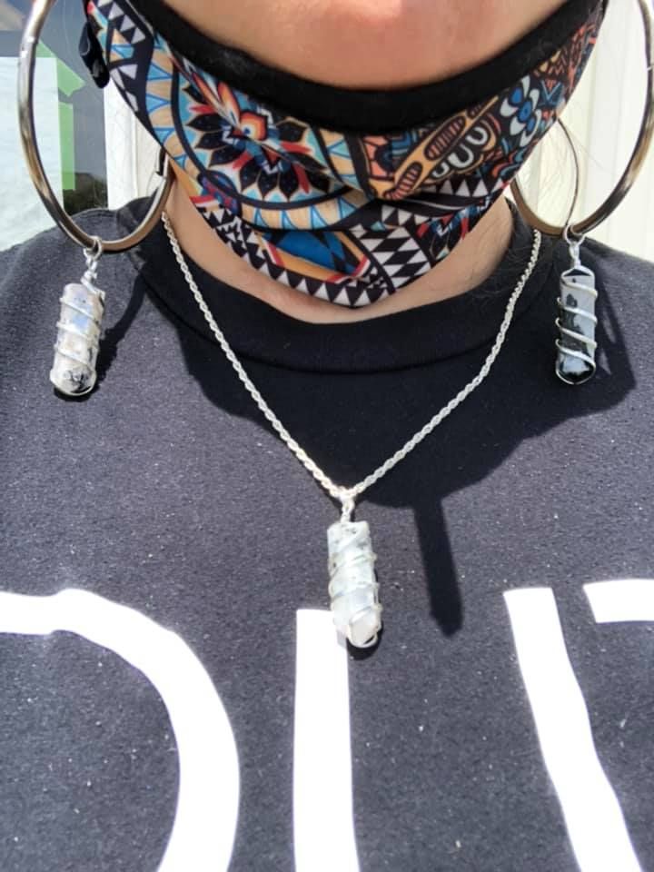Person wearing a patterned mask, large hoop earrings, and necklace with wrapped crystal pendants, black shirt in a USA shop.