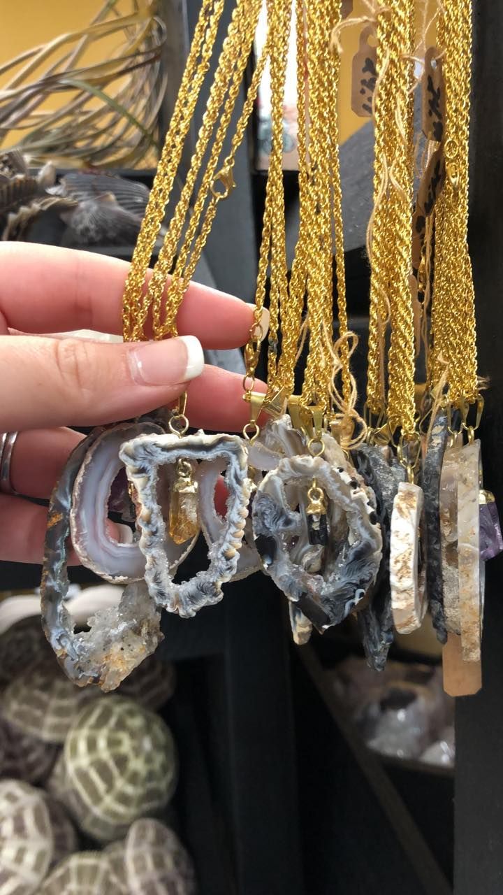 Hand holding a gold chain necklace with a gray and white geode pendant; others hang on a display in a USA shop.