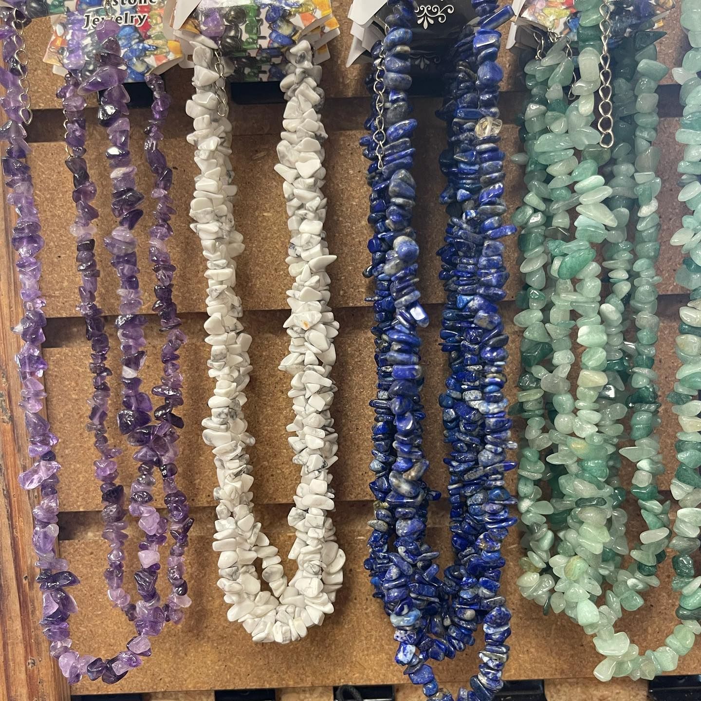 Necklaces made of amethyst, white, lapis lazuli, and green tumbled stones displayed on a wooden rack.