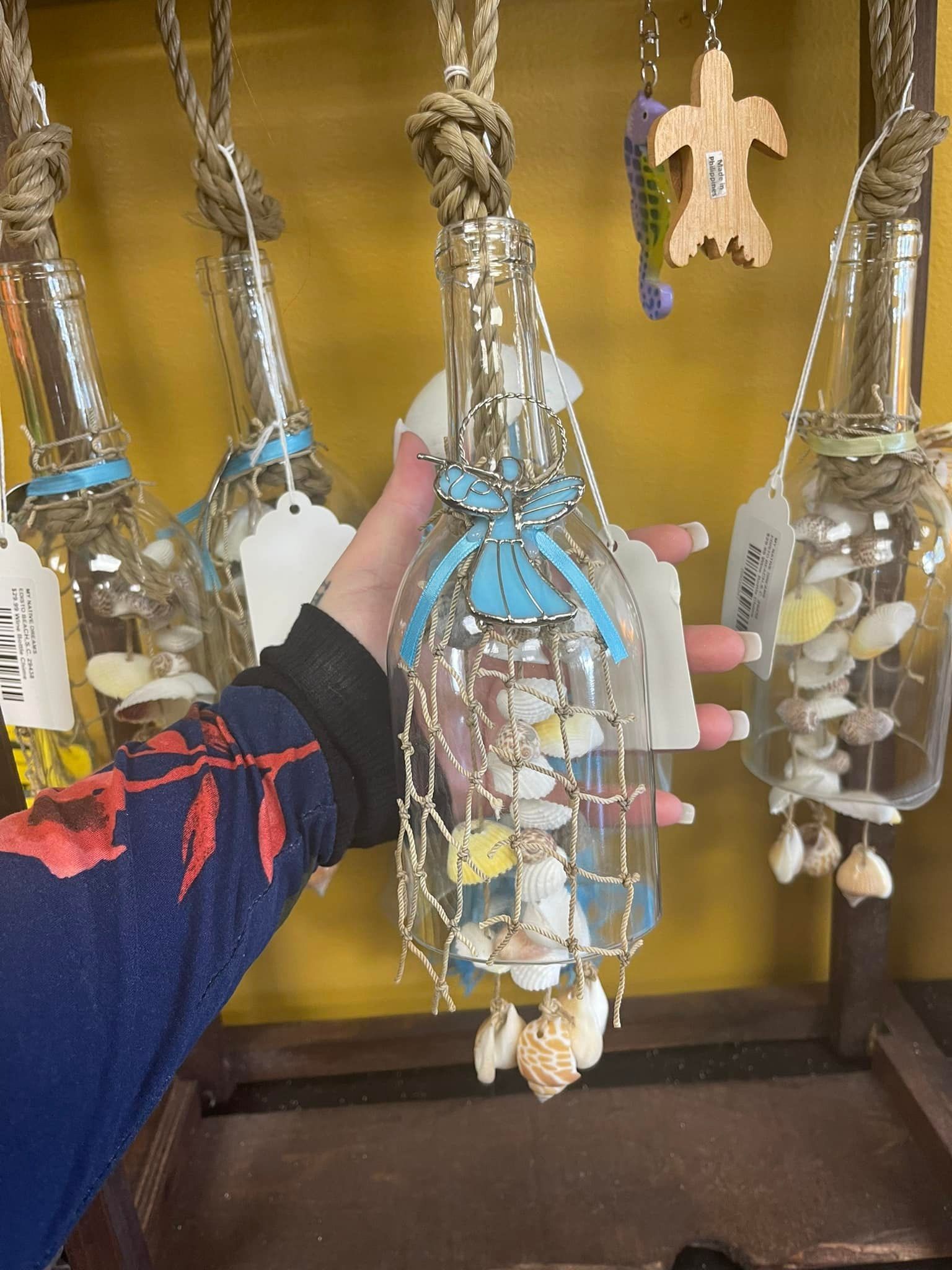 Person holding a decorative bottle filled with shells, blue ribbon, and hanging rope. Displayed in a shop.