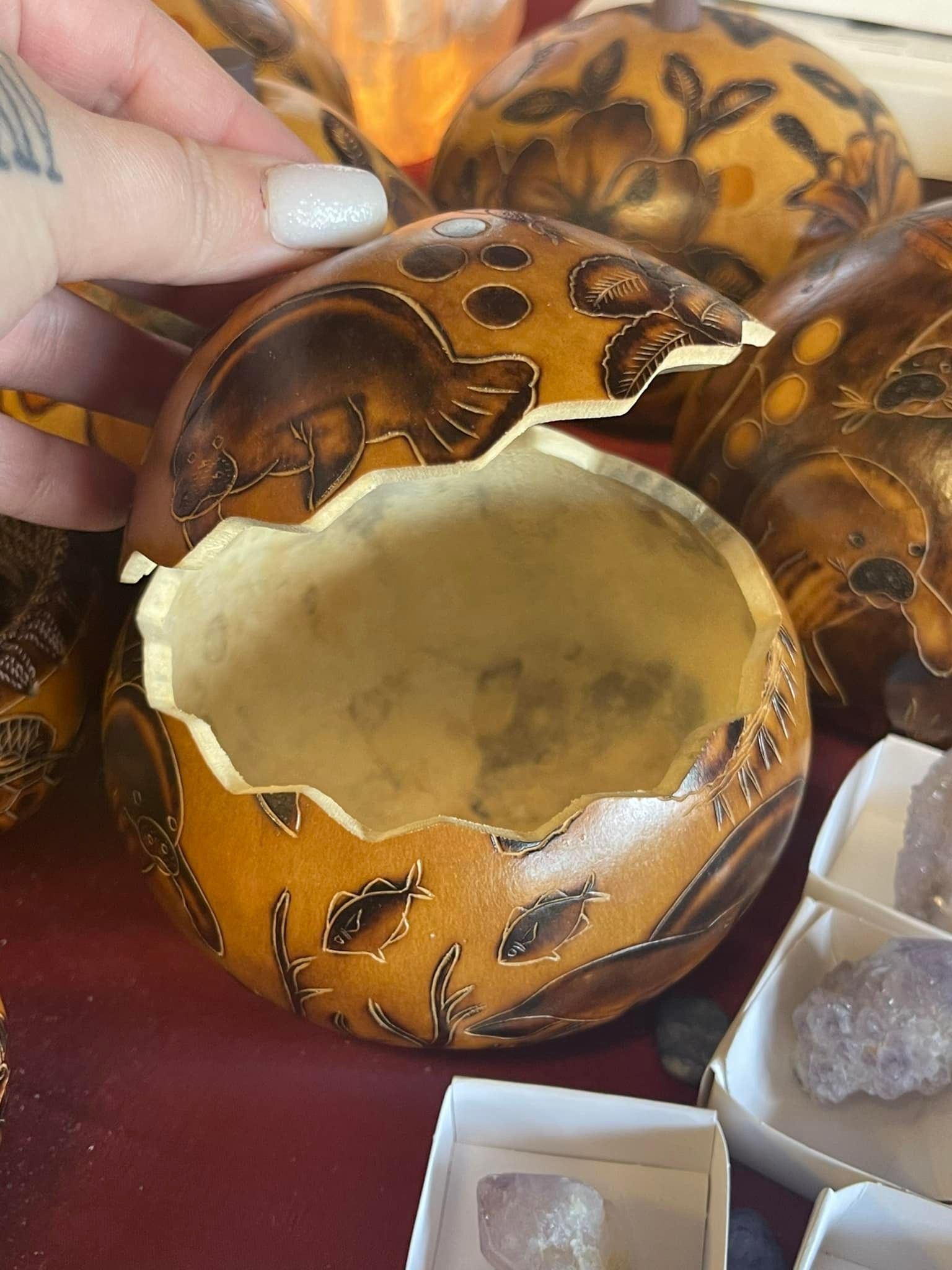 Hand holding a carved gourd bowl with leaf designs, surrounded by similar gourds and crystals.