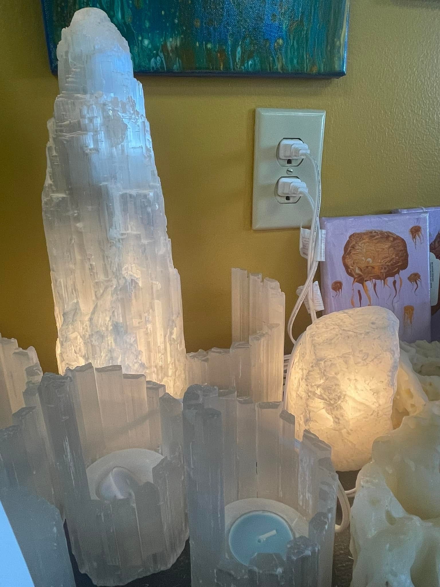 Lit selenite crystal lamps, near an electrical outlet, with a painting in the background.
