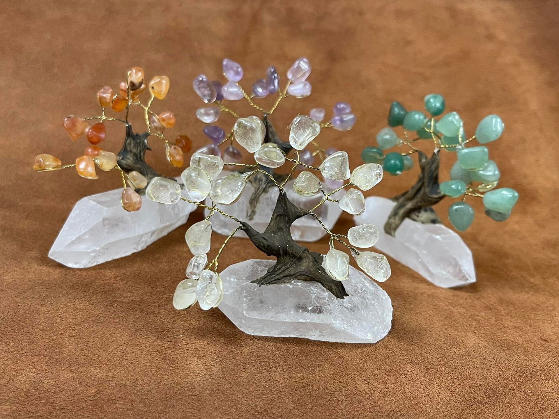 Four small crystal trees, each with colorful leaves, on clear quartz bases, set on a brown surface.