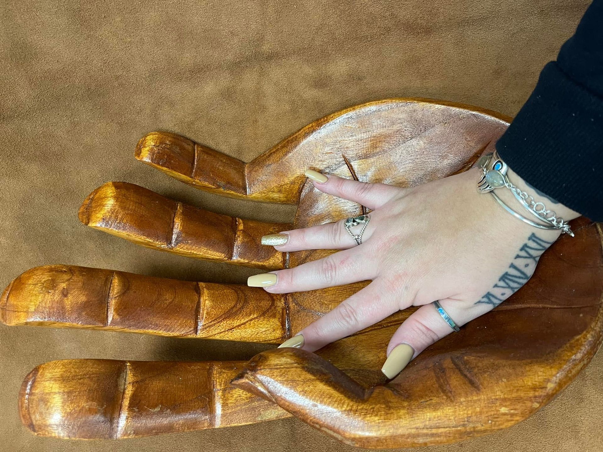 A hand with manicured nails rests on top of a large, carved wooden hand on a brown surface in a USA shop.