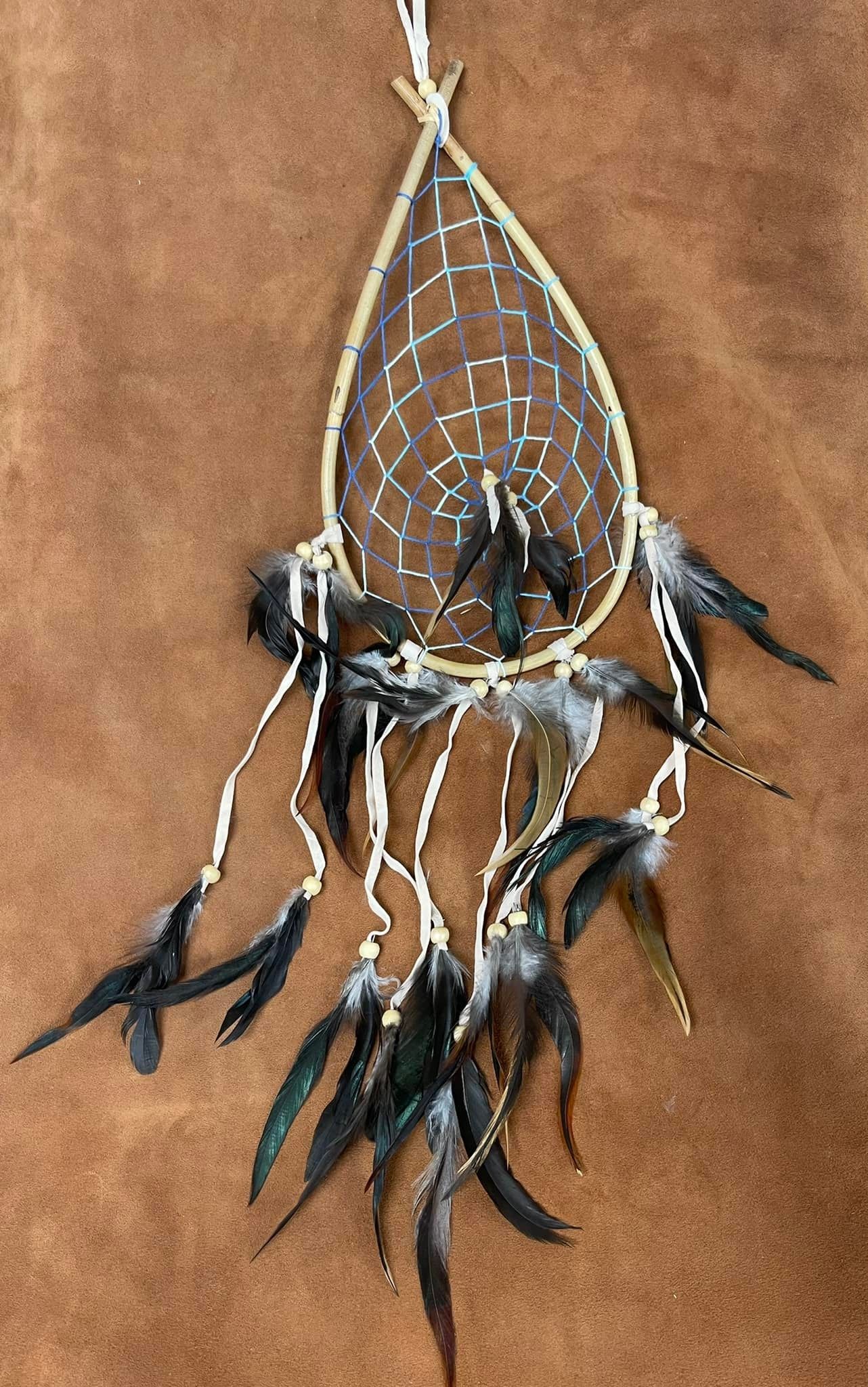 Teardrop-shaped dreamcatcher with a beaded web, black feathers, and gold accents against a brown background.
