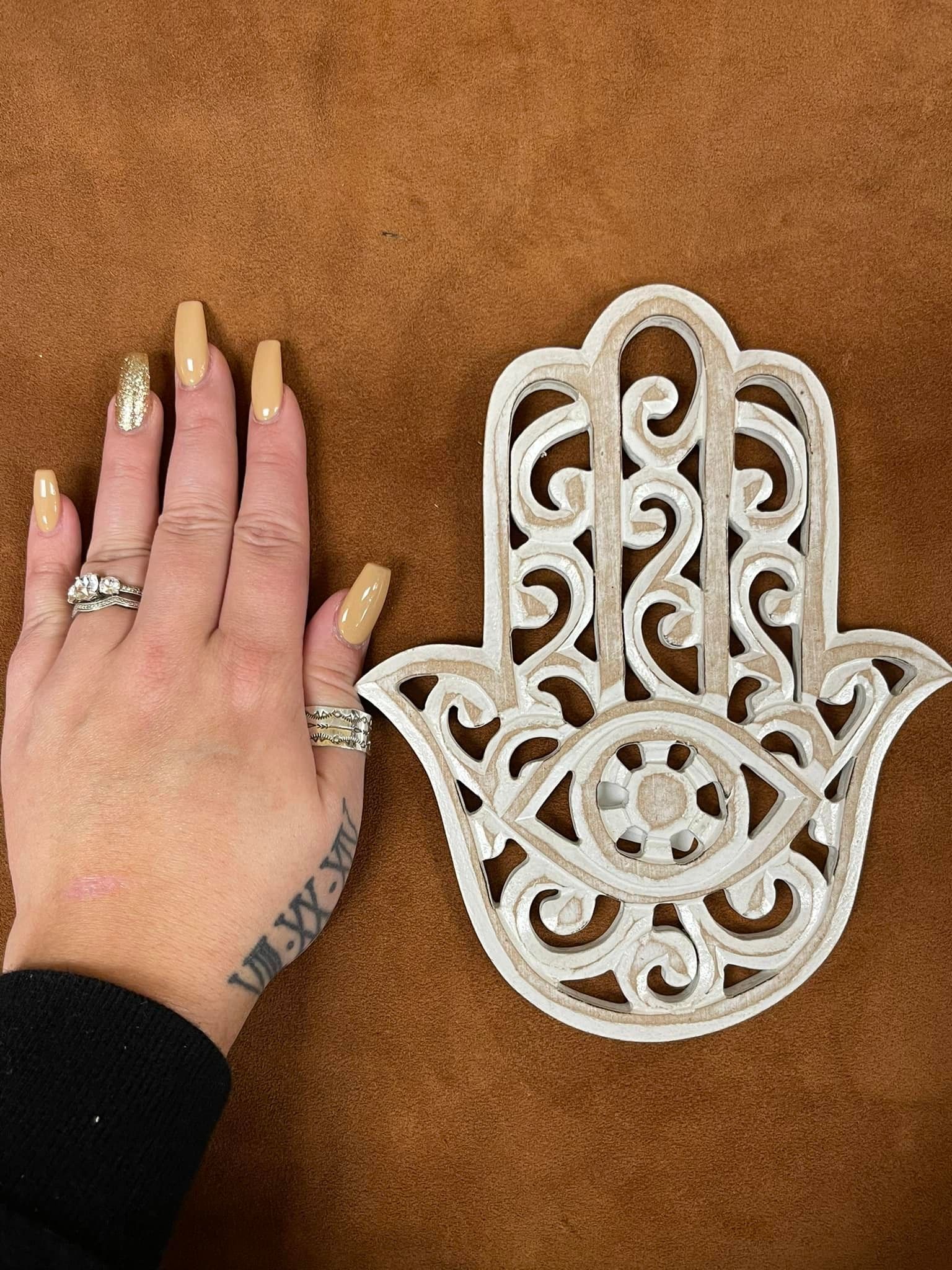 Hand with tan nails next to a white carved Hamsa hand on a brown surface.