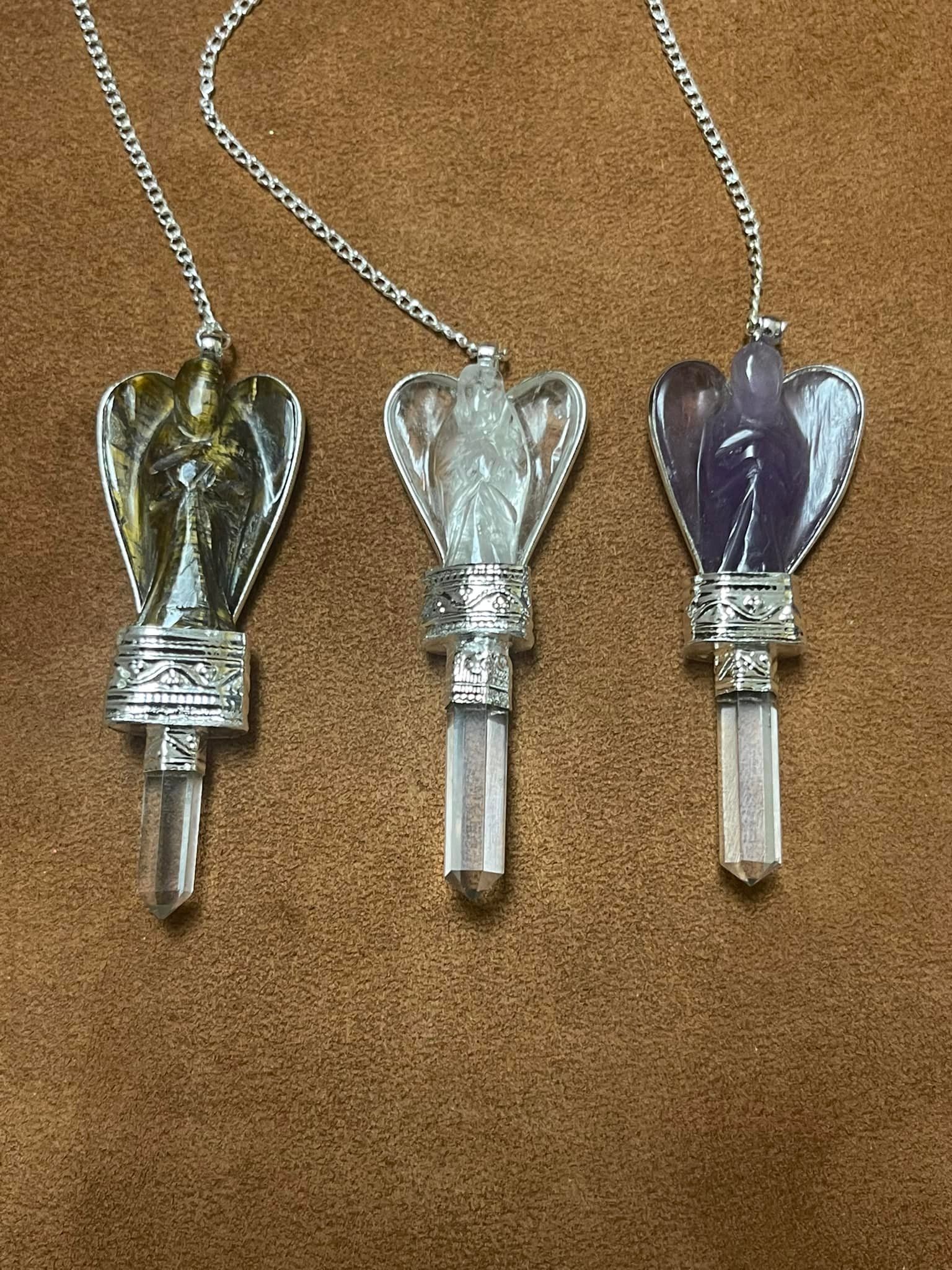 Three angel-shaped crystal pendulums: tiger's eye, clear, and amethyst, hanging on a brown background in a USA shop.
