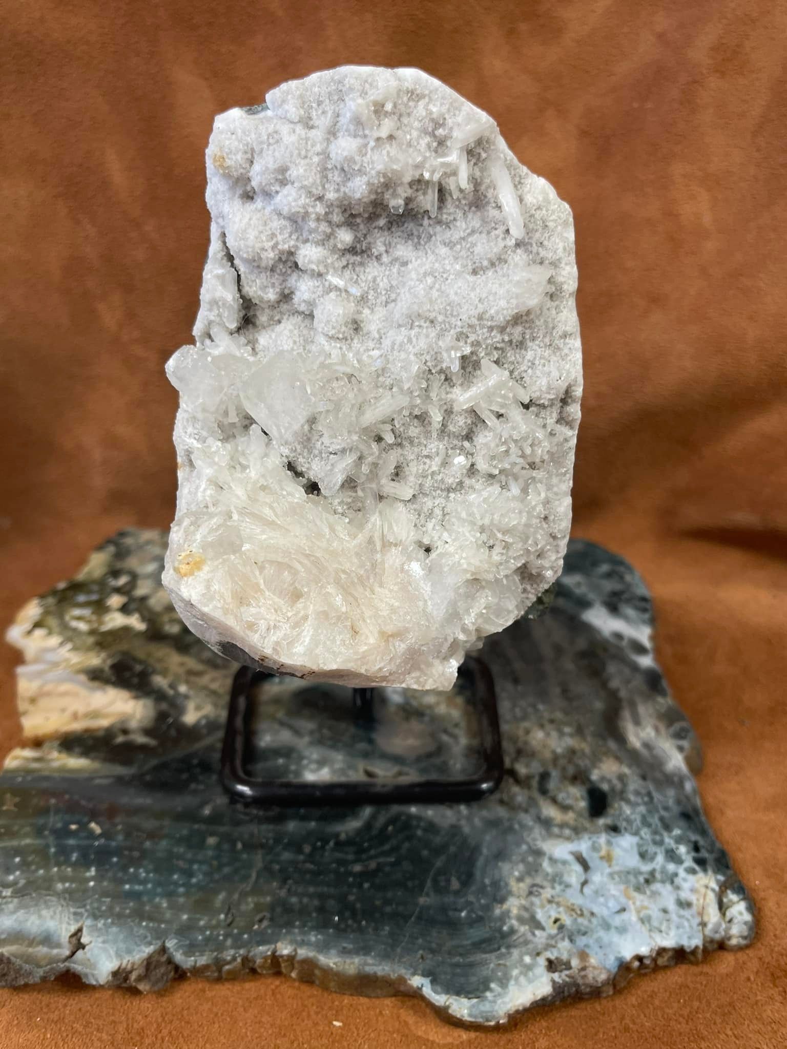A cluster of white crystals on a stand, set against a dark, patterned stone base and brown backdrop.
