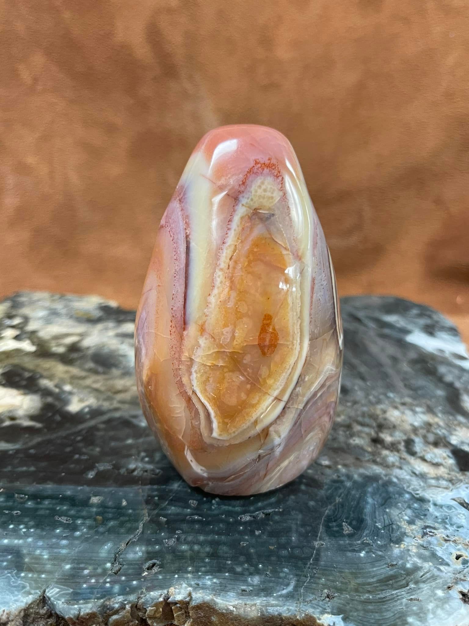 Polished orange, pink, and white agate stone, teardrop-shaped, resting on a gray rock.
