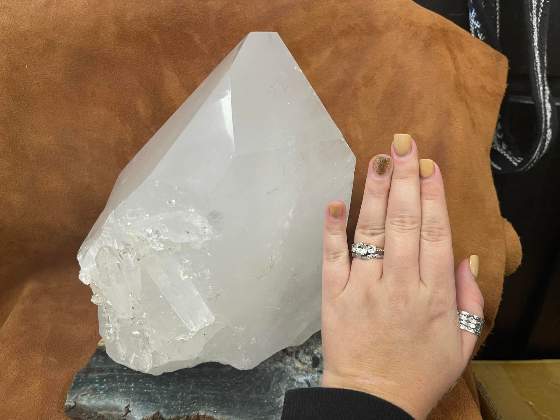 Hand next to large, clear quartz crystal. Light reflects off the crystal. Brown backdrop.