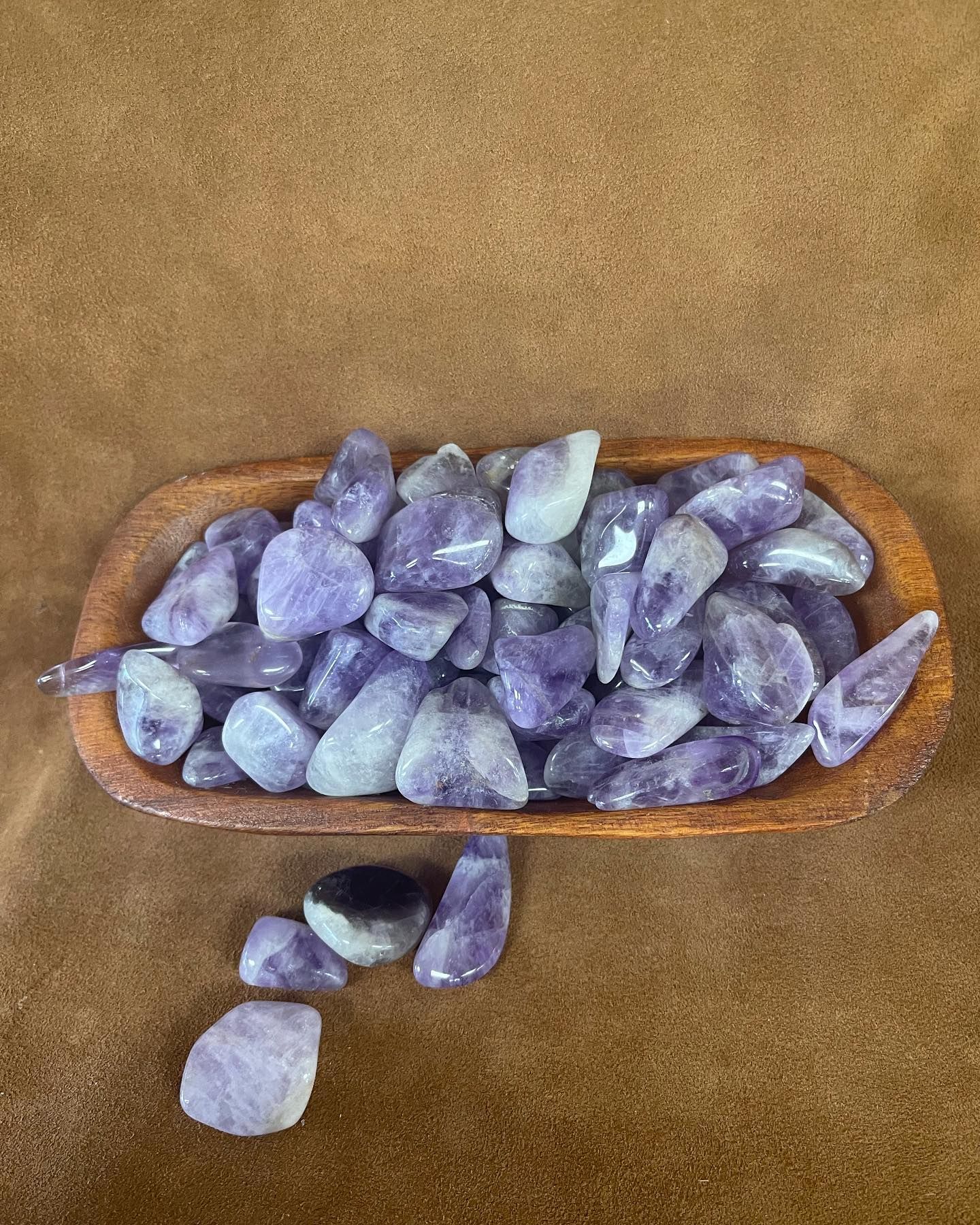 Amethyst tumbled stones in a wooden bowl on a brown surface.