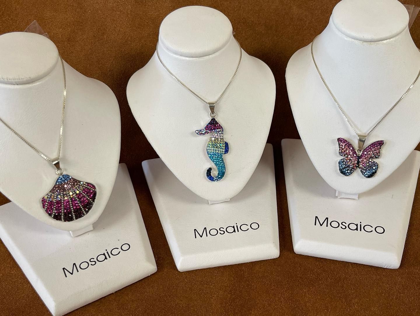Three mosaic pendant necklaces on white display busts: seashell, seahorse, and butterfly.
