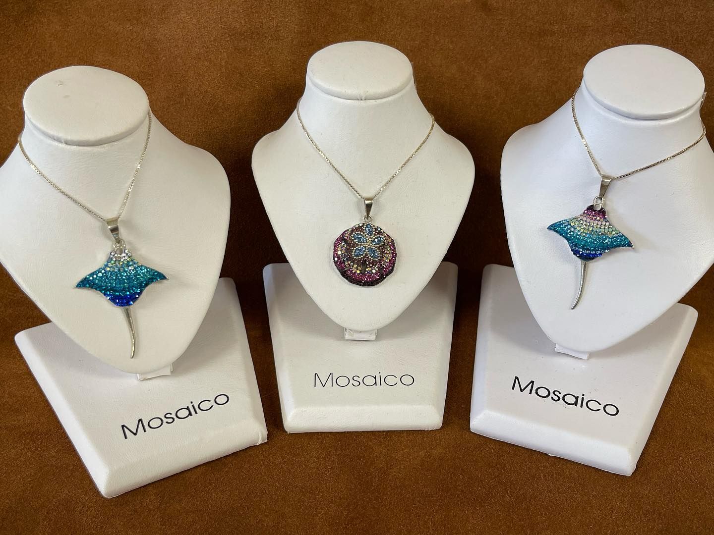 Necklaces on white stands: two blue ray pendants, one multi-colored pendant, brown background.